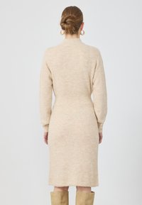 Abito a maglia beige con collo alto, maniche lunghe e vita aderente. Presenta una texture a coste e un orlo all'altezza del ginocchio.