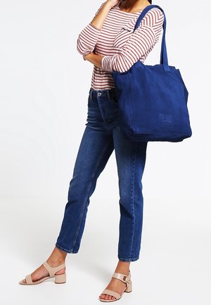 Sac fourre-tout en toile bleu marine avec deux bretelles, forme rectangulaire, surface texturée et détail de logo embossé sur le devant.