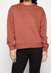 Sudadera marrón con cuello redondo, hombros caídos, puños y dobladillo de canalé. Textura suave con un detalle de costura en X en la parte delantera.