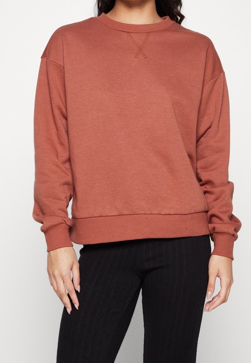 Sudadera marrón con cuello redondo, hombros caídos, puños y dobladillo de canalé. Textura suave con un detalle de costura en X en la parte delantera.