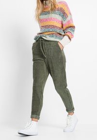 Pantalones de corduroy verde oliva con un corte ajustado y cintura con cordón. Combinados con un suéter a rayas multicolores y zapatillas altas blancas.