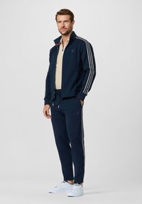 Tuta da ginnastica blu navy con righe bianche, giacca con zip e pantaloni con coulisse. Presenta un logo sulla giacca e tasche laterali sui pantaloni.