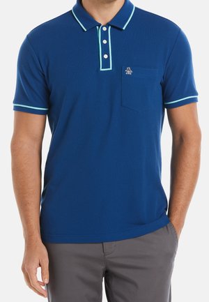 Mann trägt ein royalblaues Poloshirt mit hellblauem Besatz an Kragen und Ärmeln, kleinem Pinguin-Logo auf der Brusttasche und graue Hose.