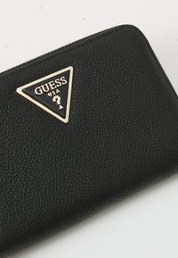 Portefeuille noir texturé avec une plaque de logo triangulaire en or, embossée avec "GUESS USA." Design de fermeture éclair lisse le long du bord.