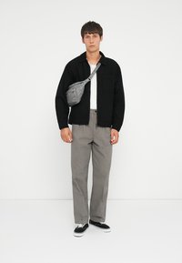 Camicia nera a bottoni sopra una maglietta bianca, abbinata a pantaloni a quadri grigi e sneakers nere. Una borsa a tracolla grigia è indossata sul petto.