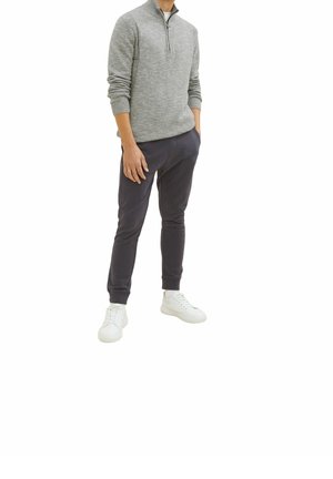 Pull en tricot gris avec col montant et fermeture éclair, associé à un pantalon bleu marine et à des baskets blanches. Texture lisse ; design ajusté.