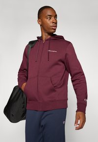 Bordeaux kleurige zip-up hoodie van een katoenmix, met een voorzak en een geborduurd logo op de borst. Vergezeld van een zwarte schoudertas.