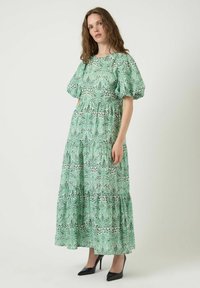 Robe longue verte à motifs floraux avec manches bouffantes, ourlet à volants et taille cintrée. Confectionnée dans un tissu léger avec un motif de feuilles répétées.