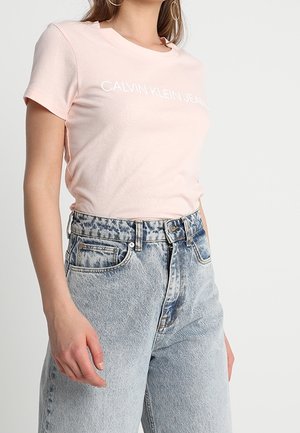 Kvinde iført en lyserød Calvin Klein Jeans t-shirt, stoppet ned i højtaljede acid-wash blå jeans med bæltestropper.