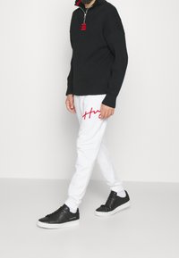 Zwarte gerebordeerde half-zip trui, witte sweatpants met rode geborduurde tekst en zwarte sneakers met witte zolen, voorzien van veters en branding.