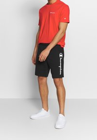 Rotes Baumwoll-T-Shirt mit Logo auf der Brust, kombiniert mit schwarzen Shorts, die ein weißes Logo an der Seite haben. Model trägt weiße Turnschuhe.