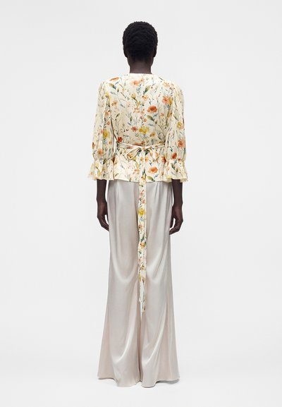 Γυναίκα με φλοράλ μακρυμάνικο μπλουζάκι με ζώνη και φαρδιά σατέν παντελόνια, στέκεται πλάτη σε μονόχρωμο φόντο.