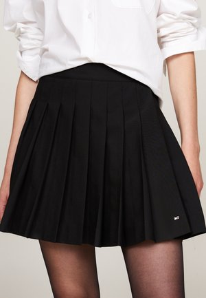 Mujer vestida con una camisa blanca abotonada metida en una falda negra plisada de talle alto con medias negras transparentes.