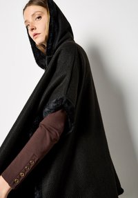 Schwarzer gewebter Poncho mit Kapuze, der an den Rändern mit weichem Kunstpelzbesatz versehen ist. Das darunterliegende Kleidungsstück ist braun und mit goldfarbenen Knöpfen akzentuiert.