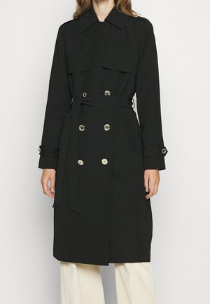 Femme portant un trench-coat noir à double boutonnage avec des boutons dorés et une ceinture à la taille, associé à un pantalon clair.
