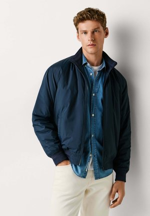 Marineblaue Bomberjacke mit Reißverschluss vorne, gerippten Bündchen und hohem Kragen, getragen über einem Jeanshemd. Verfügt über weiches, glattes Material und einen lockeren Schnitt.