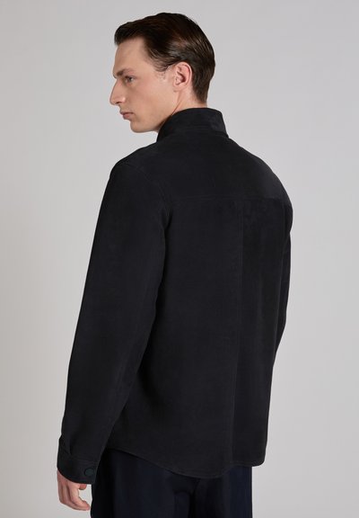 Młody mężczyzna z krótkimi brązowymi włosami, ubrany w czarną, długorękawową koszulę, ukazany z tyłu i z boku na jednolitym szarym tle.