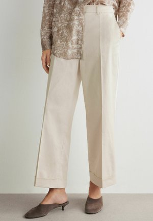 Frau trägt beige weit geschnittene Hosen mit umgeschlagenen Säumen, kombiniert mit taupefarbenen Wildleder-Mules mit niedrigem Absatz und einer gemusterten Bluse mit langen Ärmeln.