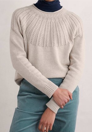 Personne portant un pull beige à torsades sur un col roulé foncé, avec un pantalon bleu-vert, les mains tenant délicatement un poignet.