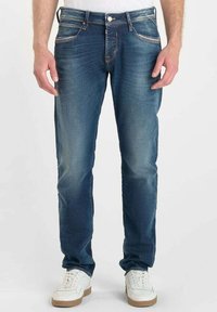 Vaqueros de mezclilla azul con un corte recto, ligero desgaste, diseño de cinco bolsillos y costuras en contraste, combinados con zapatillas blancas.