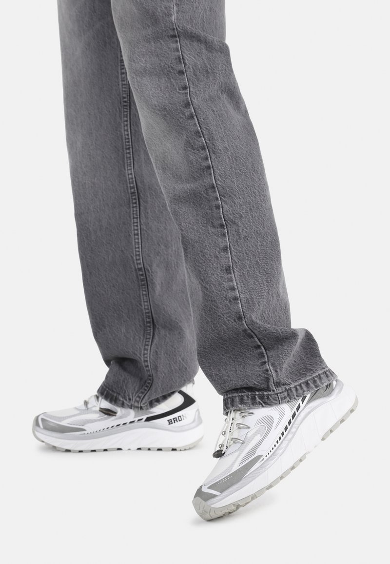 Zapatillas deportivas blancas y grises con parte superior de malla texturizada, suela gruesa y marcas prominentes. Llevadas con unos vaqueros grises de corte holgado.