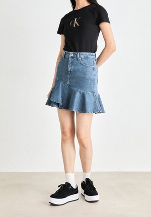 Kvinde iført sort Calvin Klein t-shirt, blå volangt denimskørt, hvide sokker og sorte platformsneakers, stående mod en ensfarvet baggrund.