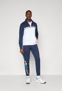 Ein Mann steht mit einem Tennisschläger in der Hand da und trägt einen navyblauen und weißen Trainingsanzug sowie weiße Sneakers vor einem schlichten hellen Hintergrund.
