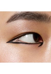 Zbliżenie na oko z odważnym czarnym eyelinerem w graficznym kształcie, gładką teksturą skóry i ciemno-brązową tęczówką na jasnym tle.