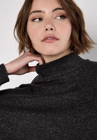 Jeune femme aux cheveux bruns courts portant un pull à col roulé gris foncé, regardant sur sa gauche avec une expression neutre.