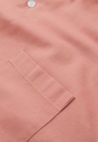 Tissu rose doux avec une poche rectangulaire cousue et un seul bouton blanc au-dessus sur un vêtement.