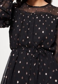 Svart klänning med genomskinliga ärmar, prydd med metalliska roséguldprickar, samlad midja med band och subtila veck vid livstycket.