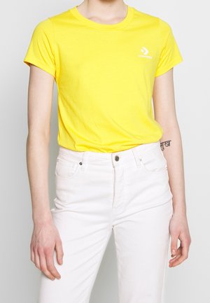 Keltainen puuvillainen t-paita, jossa on lyhyet hihat, pyöreä kaula-aukko ja valkoinen Converse-logo rintamuksessa, yhdistetty valkoisiin farkkuihin.