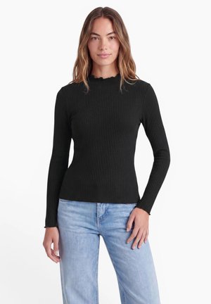 Ung kvinde med langt hår iført en sort ribbet langærmet top og lyseblå jeans, der står foran en hvid baggrund.