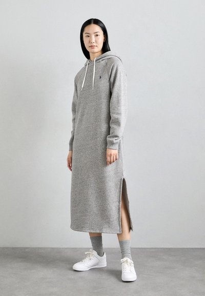 Grijze hoodie-jurk met lange mouwen, zijsplitten en een trekkoordkap. Draagt bij witte sneakers en grijze enkelsokken. Zachte stoftextuur.