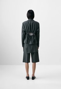Dark green jacket with a drawstring detail at the back, layered on top of matching shorts. Smooth texture and modern design.