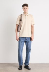 Beige pikétröja med en liten logotyp, blå jeans och svarta skor. Modellen bär en ljusbrun axelremsväska. Enkelt design och avslappnad passform.