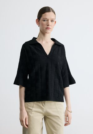 Moteris dėvi juodą tekstūruotą V formos iškirptės palaidinę su apykakle ir alkūnės ilgio rankovėmis, derinančią su smėlio spalvos kelnėmis, stovinti priešais paprastą šviesų foną.