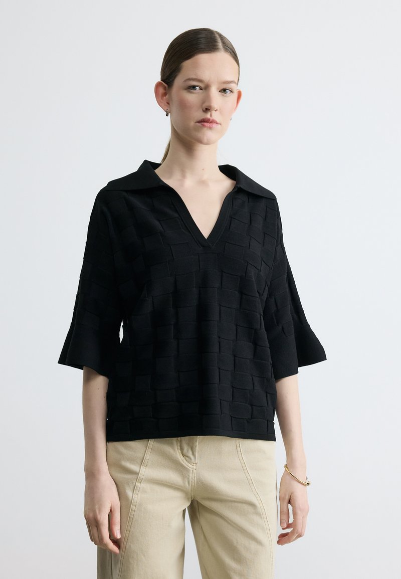 Femme portant un haut noir texturé à col en V avec col et manches mi-longues, associé à un pantalon beige, sur un fond clair uni.