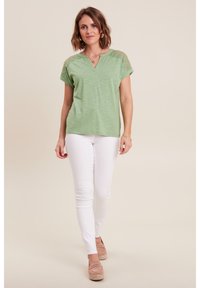 Grünes Kurzarmoberteil mit V-Ausschnitt und Spitzenakzenten an den Schultern, kombiniert mit weißen, figurbetonten Hosen und hellbraunen Espadrilles.