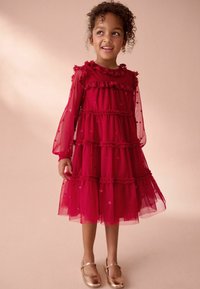 Ragazzina con capelli ricci che indossa un vestito rosso a maniche lunghe e a balze, con scarpe Mary Jane dorate, sorridente e che guarda di lato.