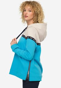 Sweat à capuche avec un design coloré : corps turquoise, capuche beige, bande noire avec un motif et ouverture latérale zippée.