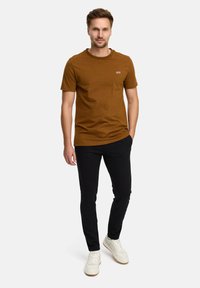 Brązowa koszulka z okrągłym dekoltem z białym napisem "JEFF", noszona z czarnymi spodniami w slim fit i białymi trampkami. Gładki materiał, luźny krój, minimalistyczny design.