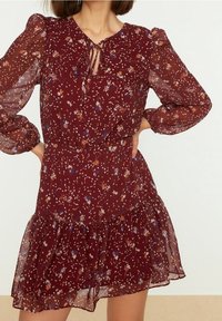 Abito floreale bordeaux con maniche lunghe a sbuffo, orlo arricciato, scollo a v e cordoncino. Presenta piccoli motivi floreali multicolori.