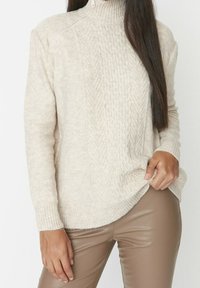 Hellbeiger gestrickter Rollkragenpullover mit Zopfmuster und gerippten Bündchen, kombiniert mit enganliegenden braunen Kunstlederhosen.