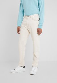 Vaqueros beige de corte recto, fabricados en denim. Presentan un diseño clásico de cinco bolsillos y herrajes metálicos. Combinados con un sudadera azul claro y zapatillas blancas.
