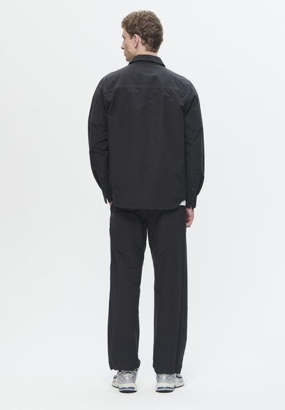 Schwarzes Langarmshirt mit Knopfmanschetten, kombiniert mit locker sitzenden schwarzen Hosen und silbernen Sportschuhen, von hinten betrachtet.