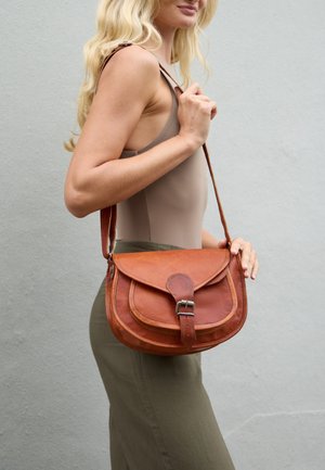 Blonde woman wearing a beige sleeveless top and green skirt carries a brown leather crossbody bag with buckle closure.