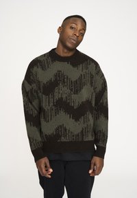 Gestrickter Pullover in Dunkelgrün und Schwarz mit Zickzackmuster, Rundhalsausschnitt und überschnittenen Schultern.