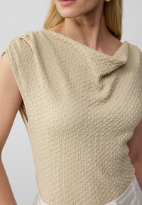 Beige top met een textuur en geweven uitstraling. Heeft een gedrapeerde halslijn en is mouwloos. Glad materiaal met een lichte rek.
