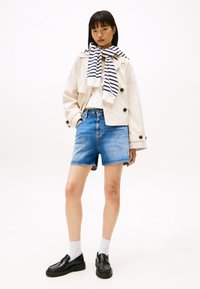 Beige cropped takki, valkoinen paita, siniset farkkushortsit, mustat loafers-kengät, yhdistettynä laivastonsiniseen ja valkoiseen raidalliseen huiviin. Lyhyet mustat hiukset, seisova asento.